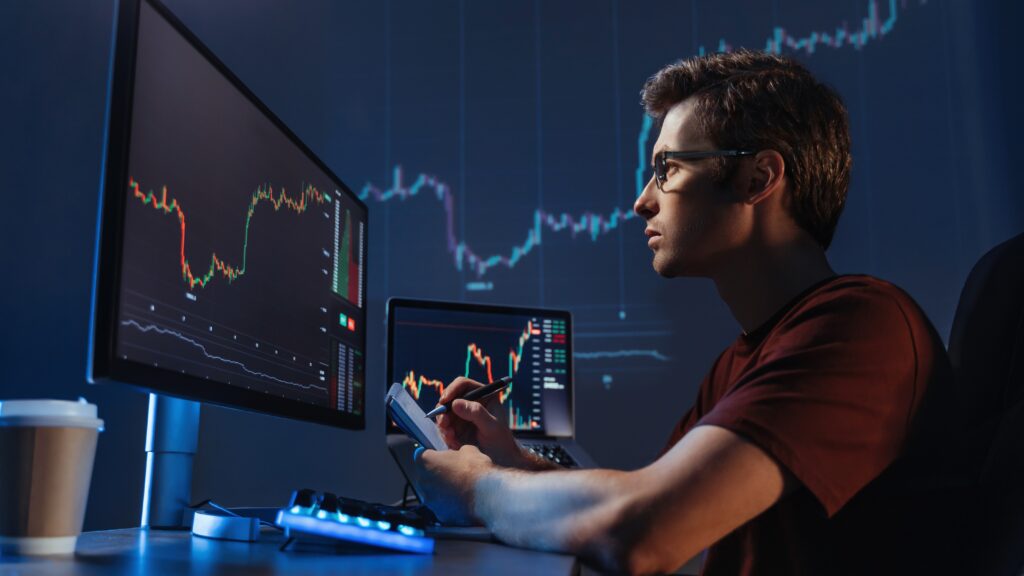 A forex trader analyzes multiple charts on computer screens, demonstrating effective risk management strategies in a dimly lit trading room. This image illustrates the importance of continuous monitoring and strategic planning in long-term survival as a forex trader. Hosted by VT Markets, a forex CFDs brokerage, for the article titled 'Risk management: Your guide to long-term survival as a forex trader.