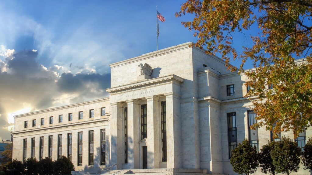 Photo of the Federal Reserve building in Washington D.C., with an American flag flying above and autumn trees in the foreground. The building is illuminated by sunlight breaking through the clouds, highlighting its architectural features. Image hosted by VT Markets, a forex CFDs brokerage.