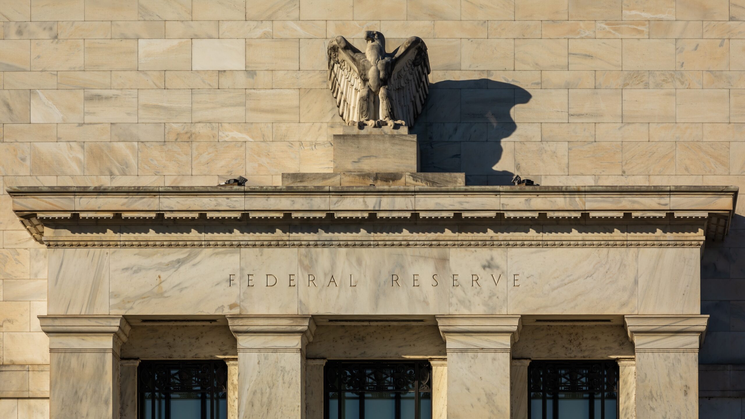 The image depicts the facade of the Federal Reserve building. The building's architectural style is classical, with a prominent stone eagle sculpture centered above the entrance. Below the eagle, the words "Federal Reserve" are engraved in the stone. The facade is composed of large stone blocks, giving it a robust and monumental appearance. The image is taken in daylight, highlighting the texture and details of the stonework.