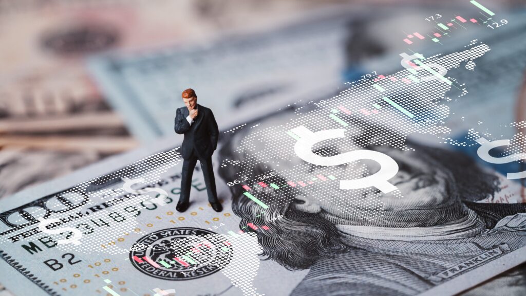 Miniature figurine of a businessman standing thoughtfully on a US dollar bill with a digital overlay of financial charts and dollar symbols, representing financial markets and trading concepts