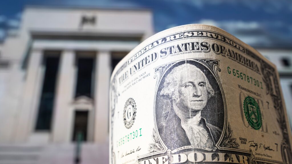 The image features a close-up of a rolled U.S. dollar bill with a portrait of George Washington prominently displayed. In the background, there is a blurred view of the Federal Reserve building, which adds a sense of depth and context to the image. The composition suggests a focus on the relationship between the U.S. dollar and the Federal Reserve. The overall tone of the image is one of economic significance, highlighting the role of the Federal Reserve in the value and stability of the U.S. currency.