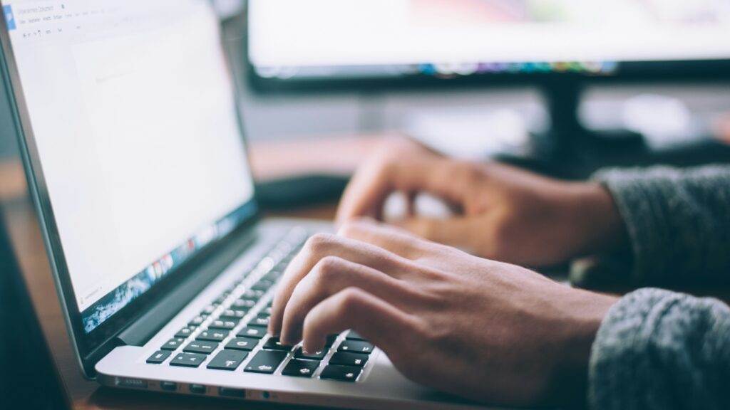 Close-up of hands typing on a laptop keyboard with a blurred screen in the background, indicative of a person working or studying.