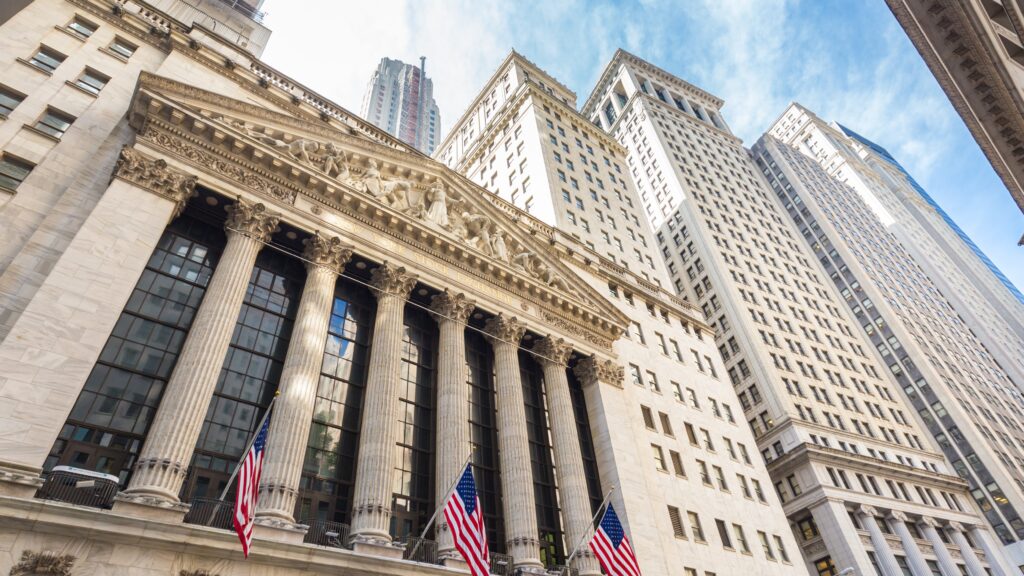 The image depicts the front facade of the New York Stock Exchange (NYSE) building, located on Wall Street in New York City. The grand architecture features tall columns and intricate carvings, with American flags displayed prominently. The sky above is clear, and the surrounding tall buildings add to the iconic financial district atmosphere.