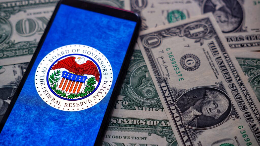 A smartphone displaying the logo of the Board of Governors of the Federal Reserve System lies on a spread of US dollar bills. This image highlights the relationship between the Federal Reserve and the US currency, relevant for articles on monetary policy, financial markets, and economic updates. Ideal for content related to forex trading, economic news, and financial analysis.